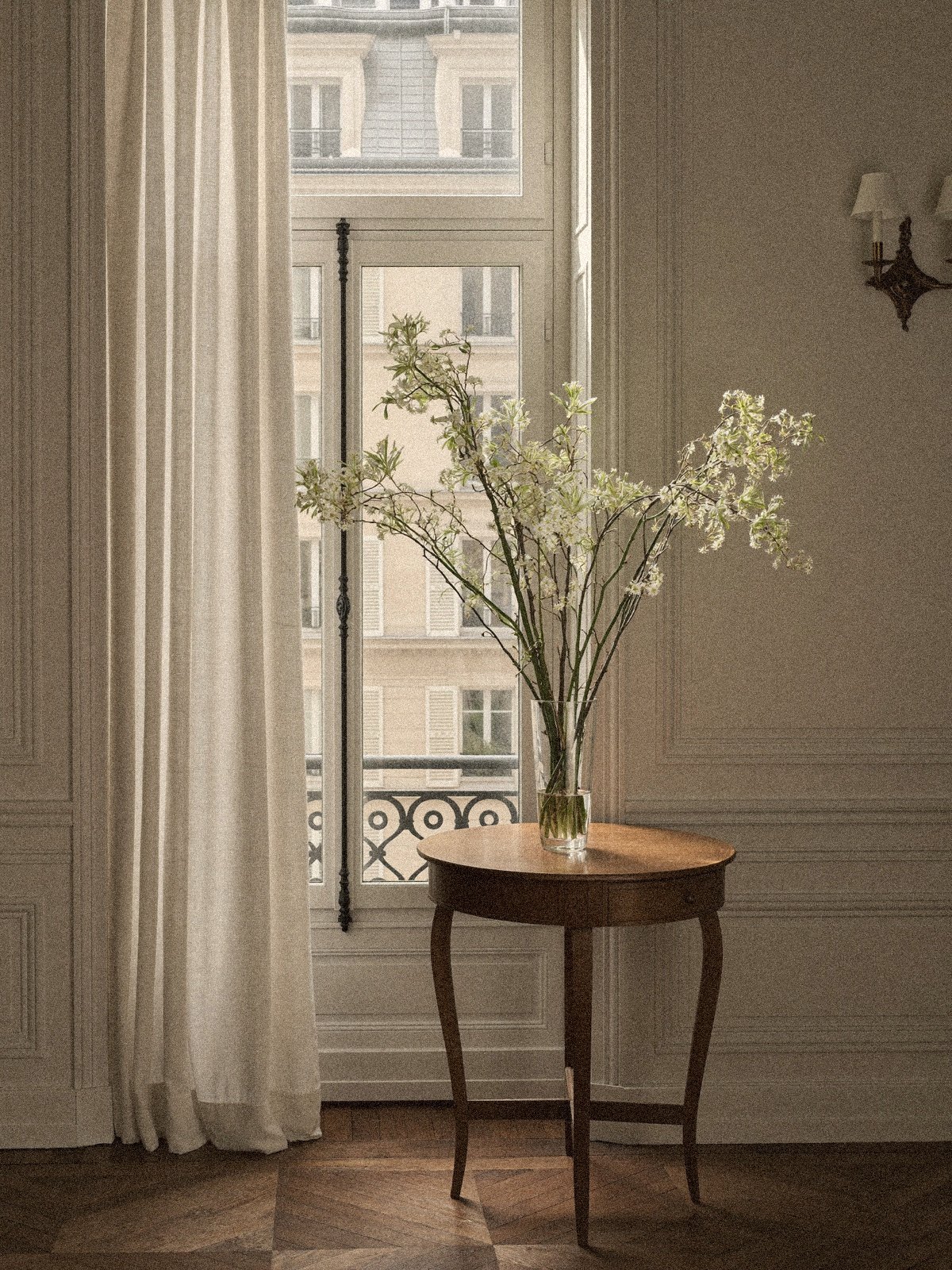 White blossom branches in a glass vase on a round wooden table beside a tall French window